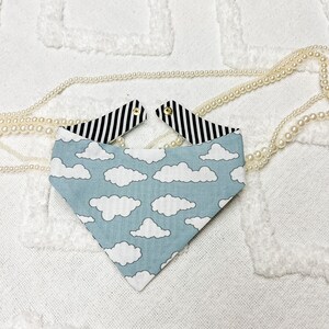 May include: A reversible pet bandana with a cloud pattern on a light blue background. The reverse side features black and white stripes. The bandana is secured with gold-colored snaps.