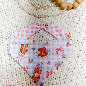 May include: A reversible pet bandana with a blue and white gingham pattern featuring illustrations of bread, pancakes, and bows. The reverse side is pink gingham. The bandana is secured with gold-colored snaps.