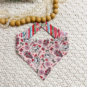 May include: A pink dog bandana with a festive Christmas pattern. The bandana features illustrations of dogs, candy canes, nutcrackers, and swirls. The top of the bandana has a striped pattern in red, green, and white. The bandana is secured with gold snaps.