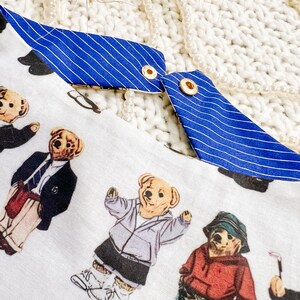 May include: White fabric featuring a pattern of teddy bears in diverse outfits, such as a blazer, hoodie, and golf attire. The fabric has blue and white striped trim with gold eyelets.