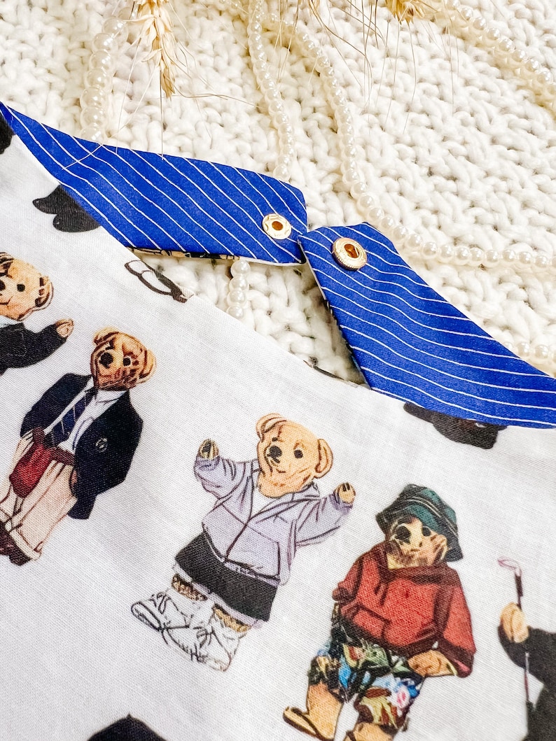 May include: White fabric featuring a pattern of teddy bears in diverse outfits, such as a blazer, hoodie, and golf attire. The fabric has blue and white striped trim with gold eyelets.