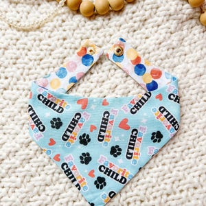 May include: A light blue pet bandana with the words "Favorite Child", paw prints, and hearts. The bandana has a colorful polka dot pattern on the adjustable straps, secured with gold snaps. The bandana is made of fabric.