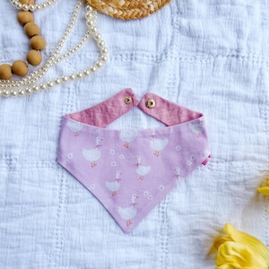 May include: A triangular pet bandana featuring a light purple fabric with a repeating pattern of white geese wearing pink bows and small white flowers. The bandana has a solid pink reverse side and two gold-colored metal snap fasteners at the top.