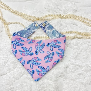 May include: A reversible pet bandana with a pink turtle pattern on one side and a blue water design on the other. The bandana has gold snap closures and is made of fabric.