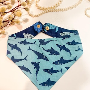 May include: A reversible pet bandana with a shark pattern on a light blue background and a turtle pattern on a navy blue background. The bandana is secured with two gold-colored snaps.
