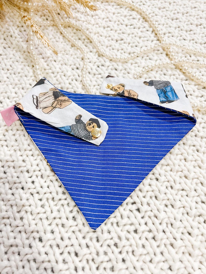 May include: A blue and white striped dog bandana with two matching collar attachments. The attachments feature a repeating pattern of teddy bears in various outfits. The bandana is made of fabric.