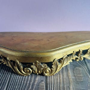 May include: A half-moon shaped, antique-style wall shelf. The shelf has a dark brown top and is supported by a gold-colored, ornate, carved base. The base features intricate scroll and leaf designs, adding a touch of elegance.