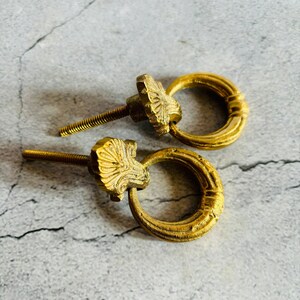 May include: Two gold-toned metal drawer pulls with ornate designs. Each pull features a ring-shaped handle and a decorative floral-shaped knob.
