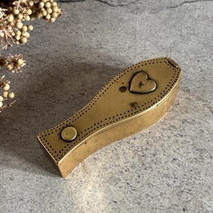 Puede incluir: Una pequeña caja de latón alargada con forma cónica similar a un ataúd. La tapa presenta una hendidura en forma de corazón, un perno circular de latón y un borde decorativo de pequeños puntos perforados a lo largo de toda la superficie.