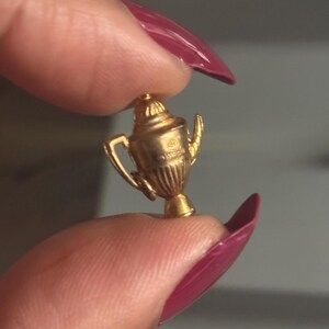 May include: A small, gold-colored trophy charm with a detailed design, including handles and a decorative top. The charm is held between two fingers with a dark background.