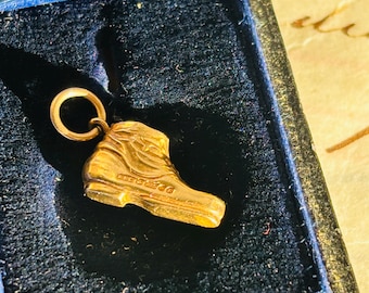Könnte beinhalten: Ein goldfarbener Stiefelanhänger mit einer Schlaufe zum Befestigen an einer Halskette oder einem Armband. Der Anhänger ist detailliert mit der Form eines Stiefels, einschließlich Schnürsenkeln und Sohle. Der Anhänger wird in einer dunkelblauen Samtbox präsentiert.