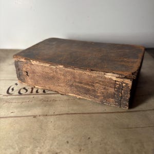Puede incluir: Un libro antiguo rectangular y desgastado, con una cubierta de cuero marrón oscuro. Los bordes de las páginas son visibles y muestran signos de envejecimiento. El libro descansa sobre una superficie con texto descolorido.