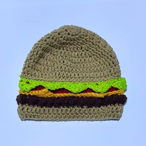 May include: A novelty crochet hat in the shape of a hamburger. The hat is made with tan yarn for the bun, green for lettuce, red for tomato, yellow for cheese, and brown for the patty. A fun, quirky accessory.