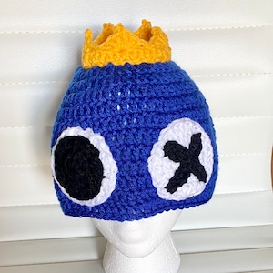 May include: A handmade blue crochet hat with a yellow crown on top. The hat features large white-rimmed eyes with black centers and a black 'X' on each side. This unique hat is perfect for costume parties or everyday wear.