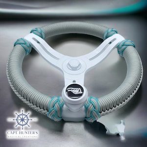 May include: A white steering wheel with a grey and teal braided rope cover. The steering wheel has a black button with a fish graphic on it. The text "CAPT. HUNTER'S NAUTICAL CREATIONS" is visible below the steering wheel.