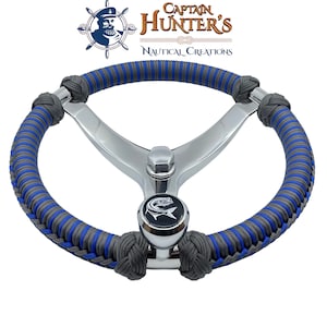 May include: A silver steering wheel with a blue and gray braided rope cover. The wheel has a three-spoke design and a fish logo on the knob. The text "Captain Hunter's Nautical Creations" is visible above the wheel.