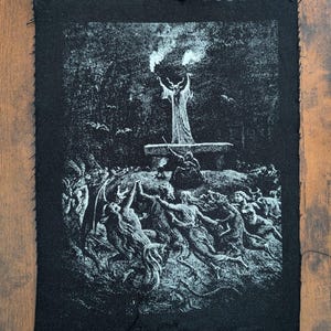 May include: A rectangular black fabric patch featuring a white screen-printed illustration of a dark ritual scene. The design depicts a central horned figure standing on an altar with raised torches, surrounded by a circle of dancing figures in a forest.