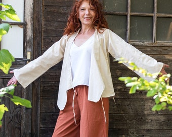 May include: A woman with long, curly red hair is wearing a cream-colored linen jacket with a white tank top underneath. She is also wearing rust-colored wide-leg pants. The woman is standing in front of a wooden wall with a window.