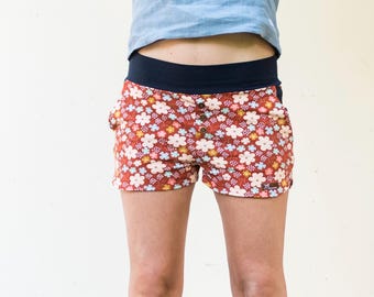 May include: Floral print shorts with a dark red background and a navy blue waistband. The shorts feature a button-up front and a colorful floral pattern with white, yellow, and pink flowers. The shorts have pockets.