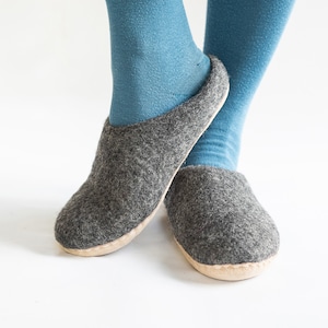 Könnte beinhalten: Ein Paar graue Filzpantoffeln mit einer hellbraunen Sohle. Die Pantoffeln werden von einer Person getragen, die blaue Socken trägt.