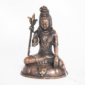 May include: Bronze-colored statue of a seated deity with intricate details. The figure has a serene expression, adorned with layered necklaces and a tall headdress. It holds a staff with a floral top and rests on a base with a carved face.