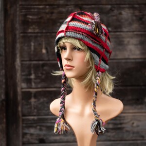 Pode incluir: Um chapéu de malha listrado vermelho, preto e cinza com longos cordões trançados. O chapéu tem uma aba para as orelhas dobrável e é usado em um manequim.