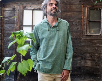May include: A man wearing a green long-sleeved shirt with a button-up collar and a brown belt. He is standing in front of a wooden building with a window.
