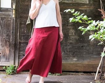 May include: A woman wearing a white camisole top and a long, flowing, red skirt. A green jacket is draped over her shoulder. The skirt appears to be made of a lightweight fabric, and the overall style is casual and comfortable.