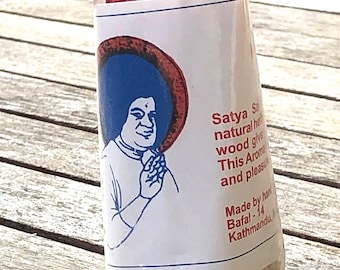 May include: A cylindrical package of incense sticks, filled with red granules at the top. The label features an illustration of a person with a blue halo and text that reads "Satya S natural he wood give This Arom and pleasur Made by han Bafal-14 Kathmandu,"