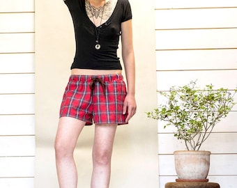 May include: A person wearing a black V-neck shirt and red plaid shorts with a drawstring. The person is wearing black lace-up boots. A small potted plant sits on a wooden stool.