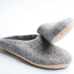 Könnte beinhalten: Ein Paar graue Filzpantoffeln mit braunem Lederbesatz. Die Pantoffeln sind umgeklappt, so dass das Innere der Pantoffel sichtbar ist.