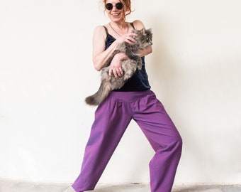 May include: A woman with red hair and sunglasses is holding a grey cat in her arms. She is wearing a purple pair of trousers and a dark blue tank top.