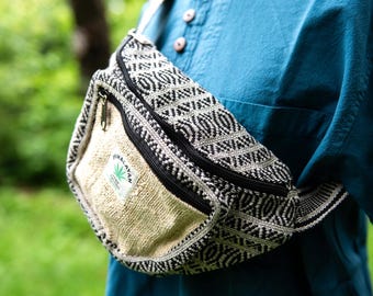 May include: A black and white patterned fanny pack with a beige front pocket. The fanny pack has a black zipper and a small tag that says "Himalayan" with a green leaf. The bag is worn over a blue shirt.
