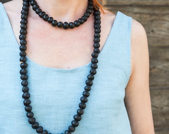 May include: A long, black beaded necklace with large, round beads. The necklace is worn over a light blue sleeveless top. The beads appear to be made of wood or a similar material. The background is a wooden wall.