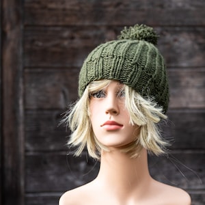 May include: A green knitted beanie with a pom pom on top. The beanie is on a mannequin head with blonde hair.