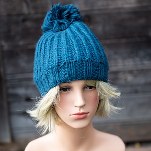 May include: A teal blue knitted beanie with a fluffy pom pom on top. The beanie is made of a soft, textured yarn.