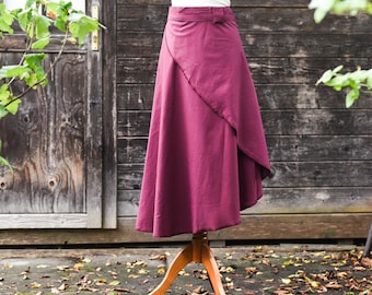 May include: A burgundy wrap skirt displayed on a wooden mannequin. The skirt has an asymmetrical hemline and a belt detail at the waist. The fabric appears to be a medium-weight material. The background features a wooden door and foliage.