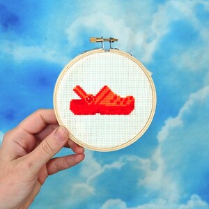 May include: A close-up of a red and orange embroidered cross-stitch design of a clog shoe on a white background. The design is framed in a wooden hoop.