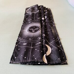 Folded black and white celestial tapestry on a bed, showing zodiac symbols, constellation lines, moon phases, and sun face print.