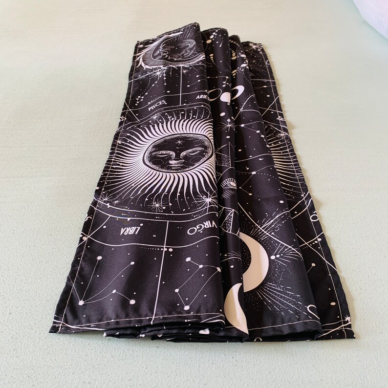Folded black and white celestial tapestry on a bed, showing zodiac symbols, constellation lines, moon phases, and sun face print.
