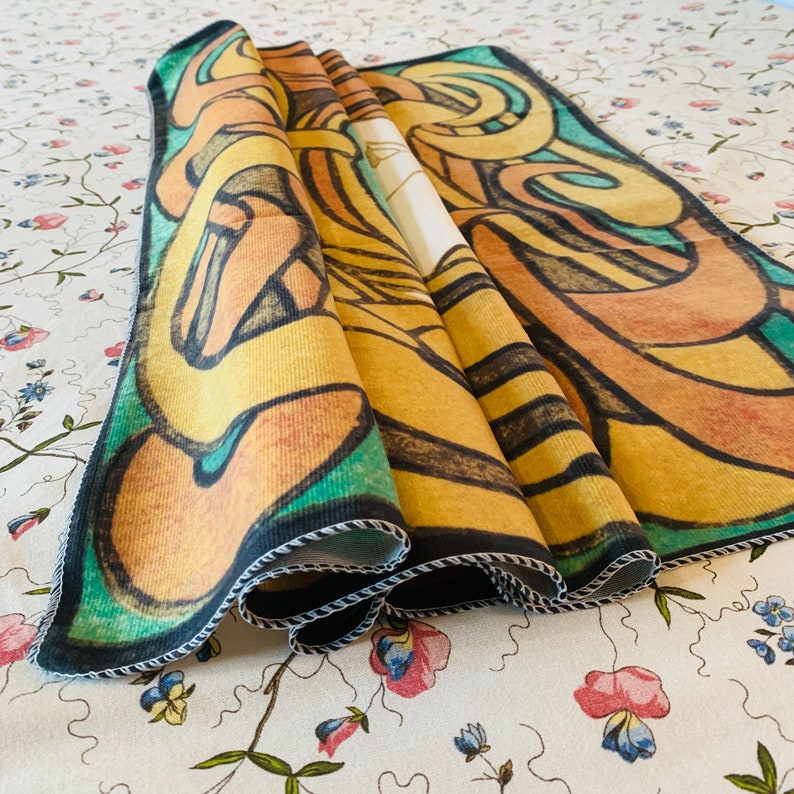 Folded brushed polyester tapestry showing the fabric texture, digitally printed yellow, orange, green and black artwork and stitched edges.