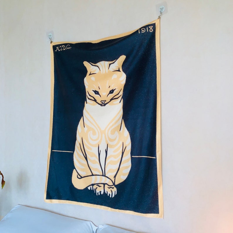 Angled view from the right of an animal art decor tapestry displayed in a cozy bedroom. The vintage 1918 illustration of a seated cat is softly illuminated by a Himalayan salt lamp resting on the bedside table.