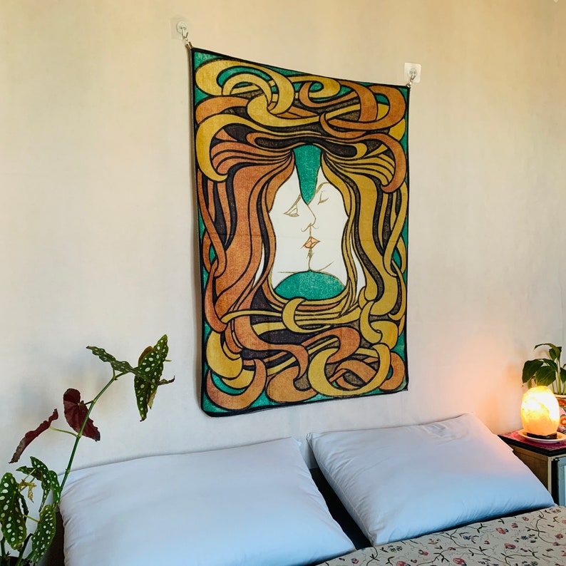 Art Nouveau The Kiss tapestry hanging above a bed, with intertwined faces, flowing yellow and orange forms, green background and black outlines.