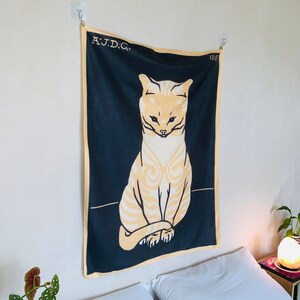 Angled view from the left of a black and yellow cat tapestry hanging over a bed with patterned sheets. A small potted begonia plant is visible in the foreground, complementing the bold vintage Dutch animal artwork.