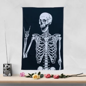May include: A black tapestry featuring a white skeleton design. The skeleton is giving a peace sign with its left hand and has a smiling face.