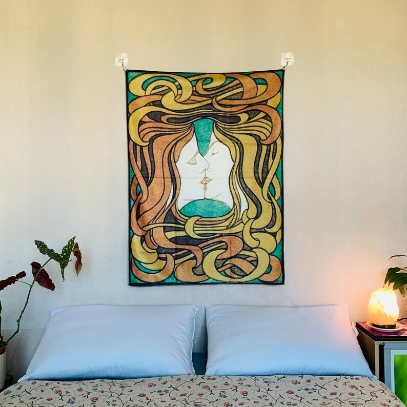 A colorful wall tapestry serving as room decor tapestry lesbian aesthetic, hung centrally over a bed with white pillows. The vintage fabric poster radiates warmth with its golden, green, and orange symmetrical kissing figures.