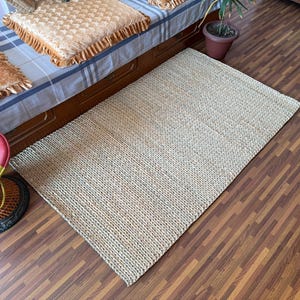 May include: A rectangular area rug crafted from natural woven fibres in a light tan, beige hue. The rug features a consistent, tight braided texture throughout its surface, lying flat on a dark wood-look floor with a visible woven construction.