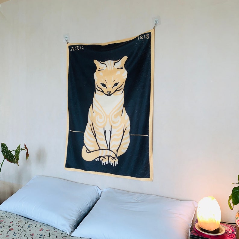 Angled view from the left of a black and yellow cat tapestry hanging over a bed with patterned sheets. A small potted begonia plant is visible in the foreground, complementing the bold vintage Dutch animal artwork.