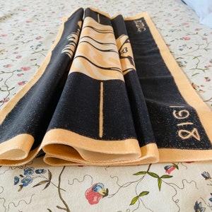 Close-up detail of the folded soft brushed polyester fabric of the Sitting Cat tapestry resting on a floral surface. The image highlights the neatly hand-stitched edges and the high-definition print quality of the black background and yellow text reading "1918."