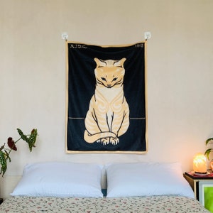 A Julie de Graag Sitting Cat tapestry hanging straight above a bed with vintage floral sheets. The black and yellow animal art wall hanging is styled next to a glowing salt lamp and a leafy green houseplant.
